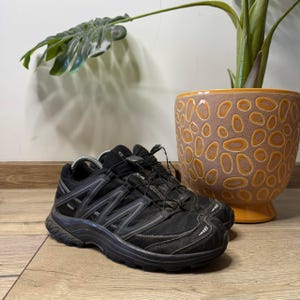 Może przedstawiać: Czarne sneakersy Salomon z cholewką z siateczki i materiału syntetycznego, systemem szybkiego sznurowania ze stoperem oraz wytrzymałą gumową podeszwą. Z boku widoczna jest nazwa marki Salomon oraz naszywka z oznaczeniem Gore-Tex.