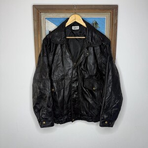 May include: A black leather jacket featuring a wide collar, a front zip fastening, and two flap pockets with gold-tone press studs. The jacket has long sleeves with press-stud cuffs and an elasticated hem, with a label reading 'Leather' inside.