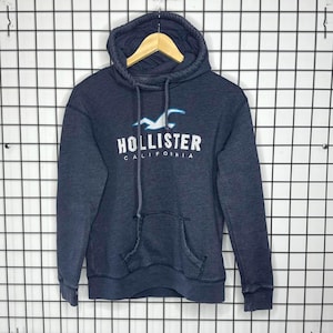 May include: A dark navy blue long-sleeve hoodie with a heathered texture. The front features the white text Hollister California and a light blue seagull logo. It includes a hood with matching drawstrings and a large front kangaroo pocket.