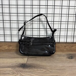 May include: A shiny black handbag with a shoulder strap. The bag has a rectangular shape with a zip closure and two decorative straps with silver buckles. The bag is set against a black grid background and wooden floor.
