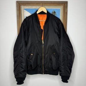 Può includere: Un giubbotto bomber nero con fodera interna arancione brillante, chiusura frontale con zip in metallo, polsini e orlo a coste e due tasche laterali con chiusura a bottone automatico. La giacca presenta maniche lunghe e un collo senza colletto.