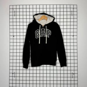 May include: Black zip-up hoodie with the white "GAP" logo across the chest. The hoodie has a white, fluffy interior lining and a drawstring hood. The hoodie is hanging on a wooden hanger against a white grid background.