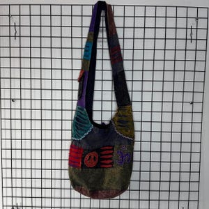 May include: A patchwork hobo bag featuring a long shoulder strap and a variety of fabric panels in shades of green, black, red, and blue. The bag displays embroidered symbols including a peace sign and an Om symbol, with distressed fabric details.