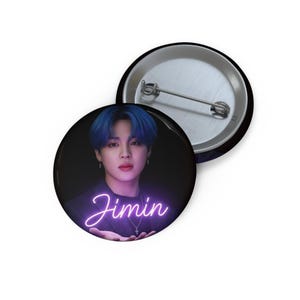 May include: A circular pin-back button featuring a portrait of a person with blue hair against a dark background. The name Jimin is written in a glowing purple neon-style script font across the lower portion of the image. The back shows a metal pin.