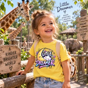 Puede incluir: Una camiseta de color amarillo brillante con un gráfico de una luna creciente sonriente, una nube y el texto "LITTLE DREAMER" en letras coloridas y estilizadas, rodeado de pequeñas estrellas y corazones, usada con vaqueros azules y mochila.