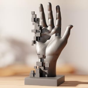 Op de afbeelding: Een grijze sculptuur van een hand op een vierkante basis. De hand lijkt te desintegreren, met secties vervangen door kleine, grijze kubussen. De sculptuur staat tegen een vage achtergrond, waardoor de gedetailleerde vorm van de hand wordt benadrukt.