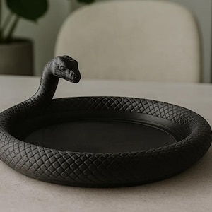 Könnte beinhalten: Ein schwarzes dekoratives Tablett mit Schlangenmuster. Das Tablett ist rund mit einem erhöhten Rand mit einem Schuppenmuster. Der Schlangenkopf ist erhoben und fügt ein einzigartiges skulpturales Element hinzu. Dieses Tablett eignet sich für Schmuck oder kleine Gegenstände.