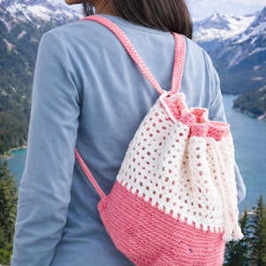 May include: A handmade crochet backpack in pink and white. The bottom half is solid pink, while the top half is white with an open weave design. The backpack has pink straps and a drawstring closure.