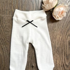 May include: White baby pants with a delicate textured pattern and a small black bow at the waist. The pants are made of a soft material and are designed for comfort. A pair of light pink roses are in the background.