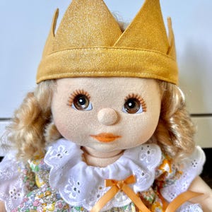 Peut inclure: Une poupée artisanale avec une couronne dorée et des cheveux blonds bouclés. La poupée a de grands yeux bruns, un petit nez et des lèvres orangées. Elle porte une robe à fleurs avec un col en dentelle blanche et un nœud orange.