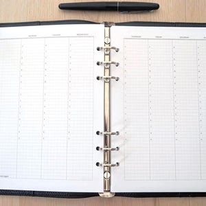 Op de afbeelding: Een open zwarte lederen map met een zilverkleurig metalen mechanisme met zes ringen en witte plannerpagina's. De pagina's hebben een rasterindeling met kolommen voor de dagen van de week. Een zwarte vulpen ligt horizontaal boven de open map.
