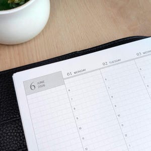 Puede incluir: Una agenda con textura de cuero negro abierta en una página que muestra el calendario de junio de 2026. El papel blanco presenta una cuadrícula con franjas horarias desde las 6 a.m., etiquetada con los días de la semana, lunes 1 y martes 2.