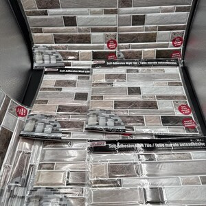May include: Multiple sheets of self-adhesive wall tiles in clear plastic packaging. Each sheet features a rectangular brick pattern in shades of brown, gray, and white. The packaging includes the text Self-Adhesive Wall Tile and 12 in / 30.4 cm.