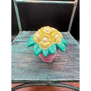 Puede incluir: Una pequeña maceta rosa contiene un ramo de flores amarillas con centros blancos y hojas verdes. Las flores están dispuestas en un racimo y las hojas son de un verde vibrante. La maceta está sobre una superficie turquesa desgastada.