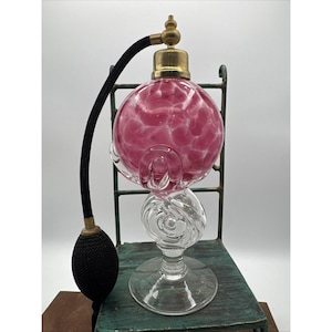 Puede incluir: Un frasco de perfume vintage con una esfera de vidrio moteado rosa y blanco, detalles metálicos dorados y un atomizador de bulbo negro. El frasco está sostenido por una base de vidrio transparente con remolinos y un marco metálico decorativo.