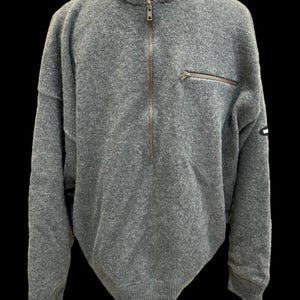 May include: A charcoal gray long-sleeve sweater featuring a mock neck collar, a central half-zip closure, and a small zippered chest pocket on the left side. The garment includes ribbed cuffs and a ribbed hem, with a small rectangular logo patch.