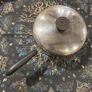 Puede incluir: Una pequeña cacerola de color plateado con tapa y asa negra. La tapa tiene un pomo negro. La cacerola está sobre una superficie oscura estampada. Utensilios de cocina.