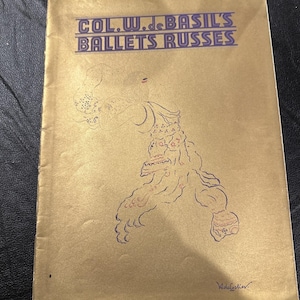 Könnte beinhalten: Ein Vintage-Programmcover mit dem Text "COL. W. de BASIL'S BALLETS RUSSES" in Lila. Das Cover ist goldfarben mit einer Skizze einer Tänzerin in Lila und Rot. Die Signatur des Künstlers befindet sich unten rechts.