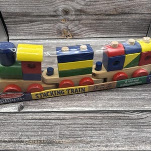 May include: A Melissa & Doug Stacking Train toy with 18 solid-wood brightly colored pieces. The train features a green engine, a yellow cab, and various blue, red, green, and yellow geometric blocks stacked on wooden bases with red wheels.