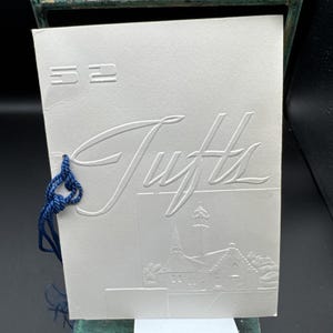 May include: A white rectangular card featuring the embossed number 52 in the top left corner and the word Tufts in large script. An embossed illustration of a building with a tall tower is at the bottom, with a blue cord tied through the left side.