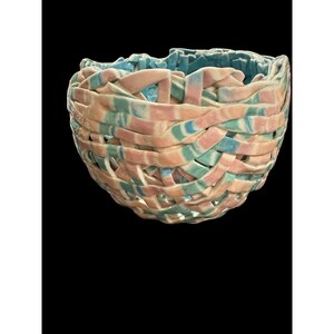 May include: A woven ceramic basket with a unique design. The basket features interwoven strips in pastel colors: pink, blue, and green. The interior of the basket is a solid light blue. The basket is set against a black background.