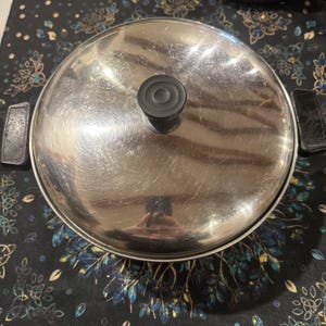 May include: A stainless steel saucepan with a matching lid. The lid has a black knob and the pan has two black handles. The pan is on a patterned surface.