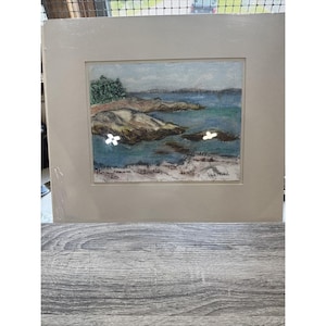 May include: A framed landscape painting depicting a coastal scene. The artwork features a blue ocean, rocky shoreline, and a light blue sky. Two white flower-like embellishments are on the painting. The painting is matted and framed in a neutral color.
