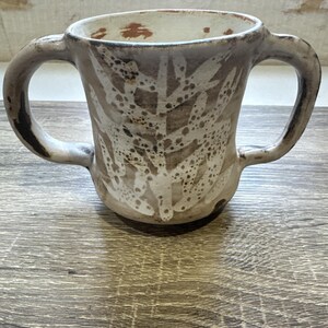 Puede incluir: Una taza de cerámica con dos asas, con un patrón de hojas en blanco sobre un fondo marrón. La taza tiene un aspecto rústico y hecho a mano con textura visible y un borde moteado. El diseño sugiere un tema inspirado en la naturaleza.