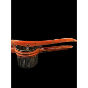 May include: Vintage orange potato ricer with a perforated basket and long handles. The kitchen utensil is made of metal and shows signs of wear, suggesting it is an antique or well-used item. The ricer is designed for mashing potatoes and other soft foods.