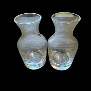 Könnte beinhalten: Zwei klare Glasvasen mit einem ausgestellten Rand und einem abgerundeten Körper. Jede Vase hat eine glatte, transparente Oberfläche mit einer subtilen horizontalen Textur im Glasmaterial und steht nebeneinander vor einem schwarzen Hintergrund.