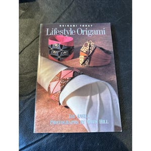 Op de afbeelding: Een boek getiteld "Lifestyle Origami" met afbeeldingen van origami servetringen. De cover toont de tekst "Origami Today" en de naam van de auteur, Jay Ansill, met foto's van Mark Hill. De servetringen zijn roze, zwart en bruin.