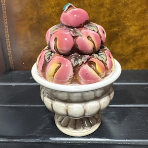 May include: A ceramic peach-shaped container with a lid. The container is a light beige color with a white rim. The lid is a stack of pink peaches with green leaves and a small green accent on top. The container is on a black surface.