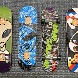 May include: Four fingerboards arranged side-by-side. The first is green and purple with an alien graphic and the text "be eve.". The second is blue and black with a geometric pattern. The third has a moon surface graphic and black wheels. The fourth is white with an alien graphic and the text "ARTO SAARI DESIGN".