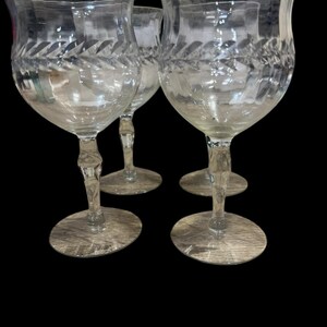 May include: A set of four clear glass goblets featuring a wide, flared rim with a subtle wavy texture. Each glass displays an etched leaf-like pattern encircling the bowl, supported by a faceted stem and a circular base, set against a black background.