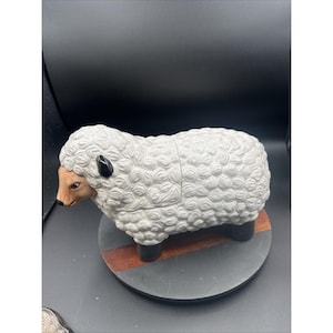May include: A ceramic sheep figurine with a textured, white woolly body. The sheep has a brown face, black ears, and black legs. It is mounted on a round, dark gray base with a wooden accent.