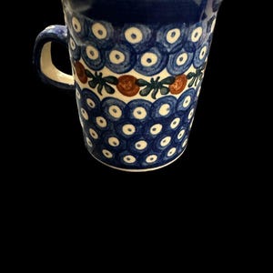 May include: A ceramic mug featuring a repeating pattern of blue and white circles, accented by a horizontal band of small orange circular shapes and green leaf-like motifs. The interior of the mug is a cream colour, and it has a single side handle.
