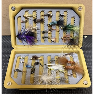 May include: A yellow plastic fly fishing box containing an assortment of artificial fishing flies. The interior features grey slotted foam inserts holding various colorful feathered and synthetic lures in purple, green, brown, and white hues.