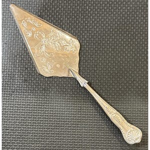 Puede incluir: Servidor de pastel plateado con un diseño floral y de cinta ornamentado. El servidor tiene una hoja triangular con un borde dentado y un mango largo y decorativo. Un utensilio clásico para servir pasteles.