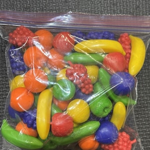 May include: A clear plastic storage bag filled with a variety of miniature plastic toy fruits, including yellow bananas, red and purple grapes, orange oranges, green apples, and green cucumbers, all featuring smooth, moulded surfaces.