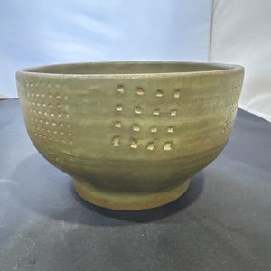 Può includere: Una ciotola in ceramica verde oliva fatta a mano con una superficie strutturata. La ciotola presenta una serie di piccole rientranze, distanziate uniformemente, disposte a griglia sull'esterno. La ciotola ha una forma arrotondata e una base leggermente rialzata.