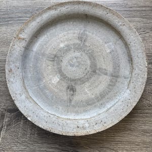 Può includere: Un piatto rotondo in ceramica con una finitura maculata grigio chiaro e beige. La superficie presenta un sottile motivo radiale che si irradia dal centro verso il bordo largo e piatto. La consistenza appare leggermente irregolare.
