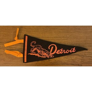 May include: Black pennant with orange detailing, displaying a tiger illustration and the word "Detroit." The pennant is triangular with an orange fabric loop for hanging. Likely a sports souvenir or memorabilia item.