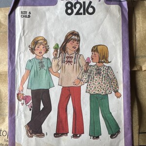 Può includere: Una busta per cartamodello Simplicity 8216 per un completo da bambino taglia 6. Il fronte presenta un'illustrazione di tre bambini che indossano top a maniche lunghe con nastri e pantaloni a zampa, con un prezzo di $1,25 stampato in alto a destra.