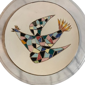May include: A round decorative plate with a cream-colored background featuring a central mosaic-style bird illustration. The bird is composed of various geometric shapes in shades of black, pink, blue, green, and yellow, outlined in gold.