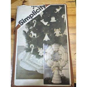 Puede incluir: Un patrón de costura Simplicity vintage con un árbol de Navidad con adornos, incluyendo ángeles, bastones de caramelo y una falda de árbol. La portada del patrón tiene un borde marrón y el logotipo de Simplicity.