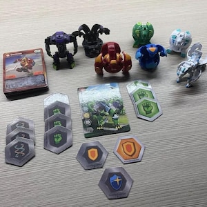Peut inclure: Une collection de jouets Bakugan comprenant six petites figurines en plastique de différentes couleurs, une pile de cartes de jeu et plusieurs tuiles hexagonales avec des symboles de poing, de bouclier et d'ADN. Les figurines sont violettes, noires, rouges, vertes, bleues et blanches.