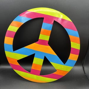 May include: A circular peace sign wall decoration featuring horizontal stripes in bright neon colours including lime green, hot pink, orange, and sky blue. The design consists of a solid outer ring with a central peace symbol cutout pattern.
