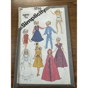 Puede incluir: Un patrón de costura Simplicity vintage, número 5356, con ilustraciones de diseños de ropa para muñecas. La portada muestra varios atuendos, incluyendo vestidos, un vestido de novia y una capa con capucha. El patrón está sobre una superficie de madera.