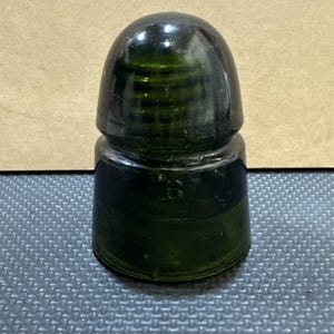 May include: A dark green glass electrical insulator with a rounded top and a cylindrical base. The object features a series of horizontal ridges around the upper section and a smooth, wide bottom edge, reflecting light on its polished surface.