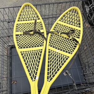 May include: Pair of bright yellow snowshoes with black straps. The snowshoes have a lattice design and are oval-shaped. The straps are attached to the snowshoes with metal buckles. The snowshoes are designed for winter outdoor activities.